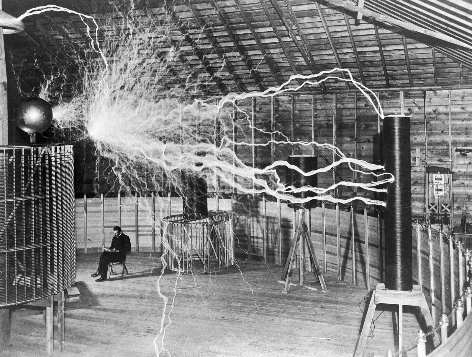 A multiple-exposure photograph of Nikola Tesla seated in the Colorado Springs laboratory, combined with a separate exposure of electrical discharges from the magnifying transmitter